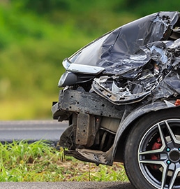 Avant droit d'une voiture noire lourdement accidentée, capot et pare-chocs déformés, garée près d'une route avec de la végétation en arrière-plan.