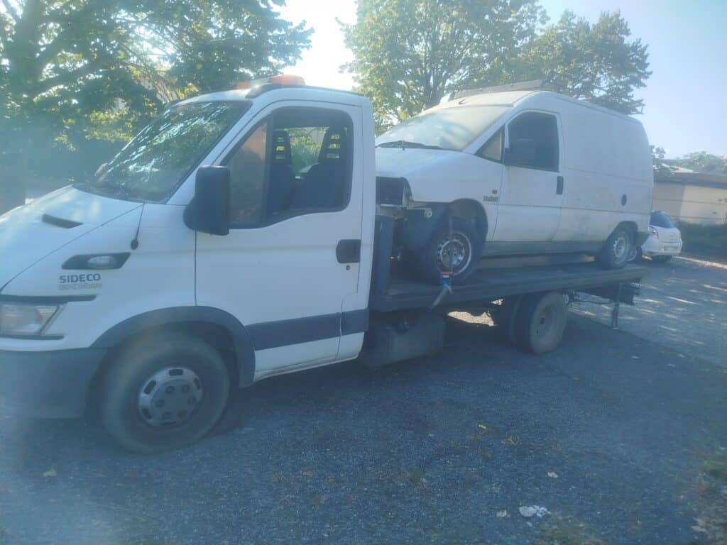 Camion plateau Iveco SIDECO pour transport de fourgonnette Camion plateau SIDECO 35 C 14 HPi transportant une fourgonnette blanche accidentée "Turbo D". Scène ensoleillée sous les arbres.