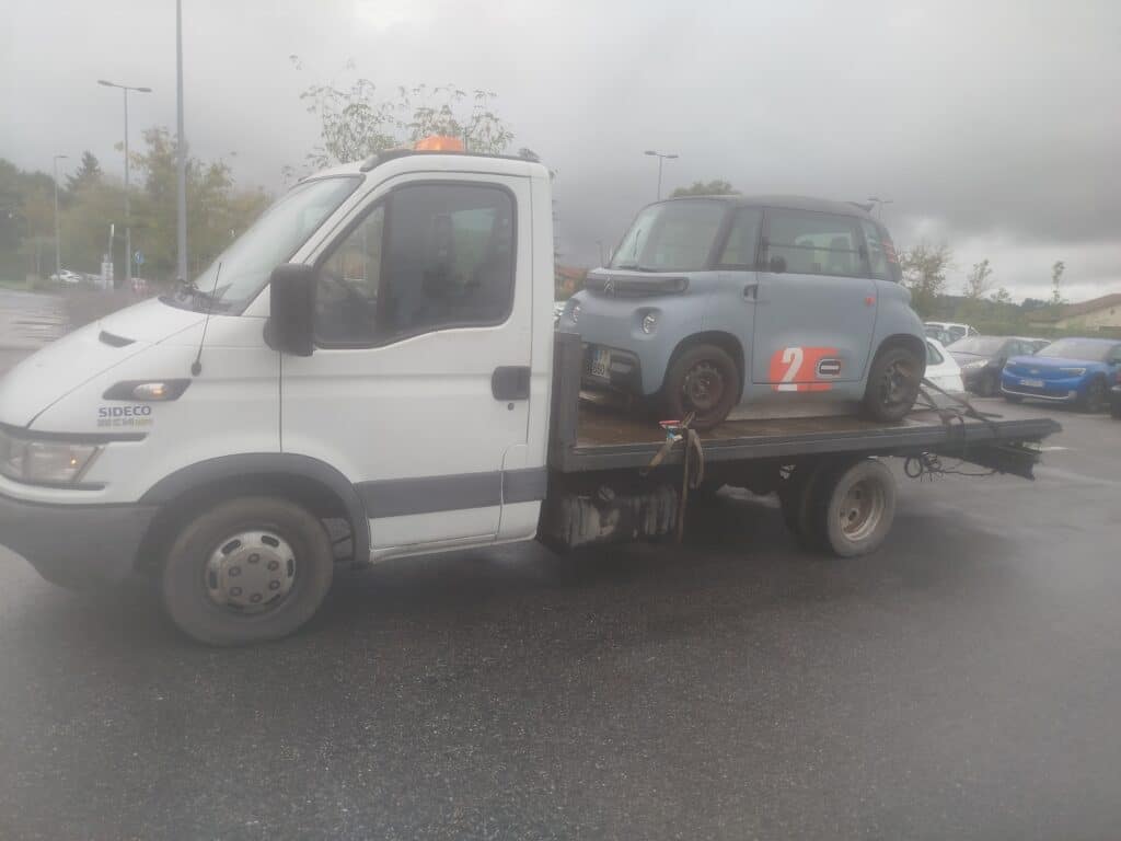 Transport d'une Citroën Ami "Race" sur dépanneuse Iveco. Dépanneuse Iveco Daily 35 C 14 HPI transporte un Citroën Ami gris de course (numéro 2) sur plateau dans un parking brumeux.
